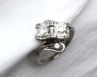 May include: A silver-toned ring featuring two prominent round-cut clear stones set side-by-side in a rectangular frame, with a third smaller round-cut clear stone positioned on a curved, polished band that sweeps beneath the main setting.