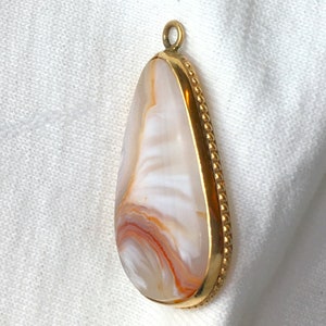 Peut inclure: Un pendentif en forme de larme avec une pierre d'agate blanche et orange sertie dans un cadre en m&eacute;tal dor&eacute;. Le cadre est bord&eacute; de perles.