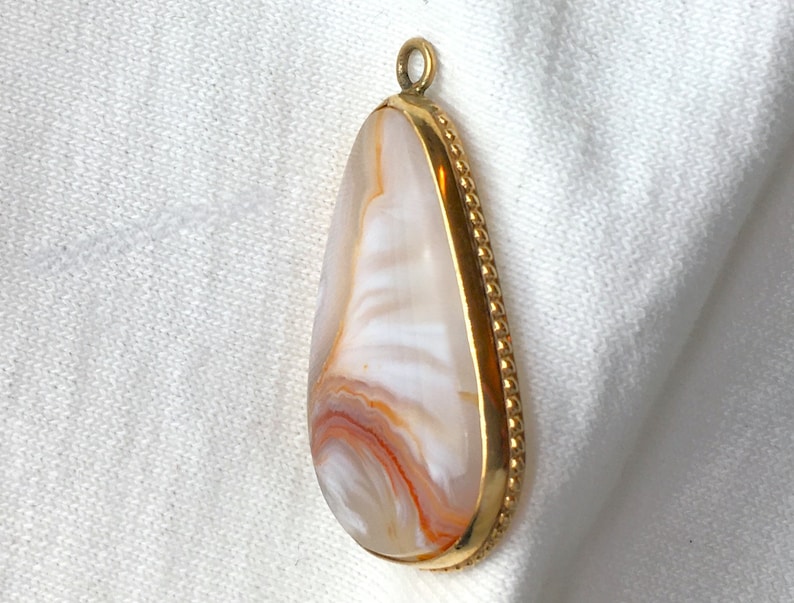 Peut inclure: Un pendentif en forme de larme avec une pierre d'agate blanche et orange sertie dans un cadre en m&eacute;tal dor&eacute;. Le cadre est bord&eacute; de perles.
