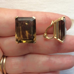 May include: A pair of rectangular, smoky-brown gemstone clip-on earrings with gold-colored metal settings. The earrings have a classic, elegant design, with a large, faceted stone in each setting. The clip-on mechanism is visible on one earring.