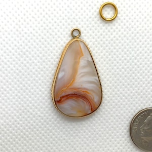 Peut inclure: Un pendentif en forme de larme avec une pierre d'agate blanche et orange sertie dans un cadre en m&eacute;tal dor&eacute;. Le pendentif a un bord perl&eacute; et une boucle pour attacher une cha&icirc;ne.