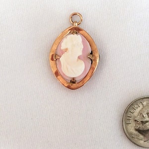 May include: A gold-toned pendant with a pink cameo featuring a woman's profile. The cameo is set in a gold-toned frame with a diamond-shaped design.