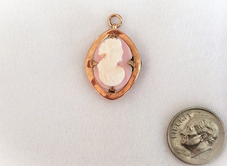 May include: A gold-toned pendant with a pink cameo featuring a woman's profile. The cameo is set in a gold-toned frame with a diamond-shaped design.