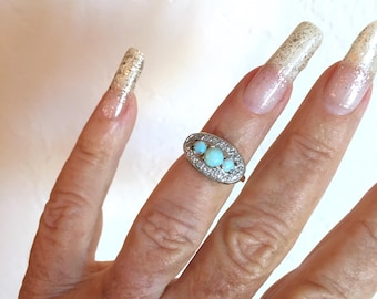May include: Oval-shaped silver ring featuring three light blue opal stones, surrounded by small clear crystals. The ring is displayed on a finger with long, glitter-covered nails. A piece of jewelry.