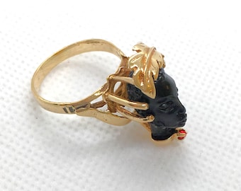 May include: A gold ring with a black cameo of a woman's face. The cameo is set in a gold frame with a gold leaf design. The ring has a red stone on the side.