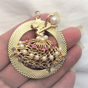 May include: A gold pendant featuring a ballerina figure with a pearl and pink and blue gemstone embellishments.
