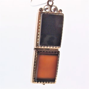May include: A gold locket with a hinged opening. The locket has two compartments, each with a different colored stone. The top compartment has a black stone, and the bottom compartment has an orange stone.