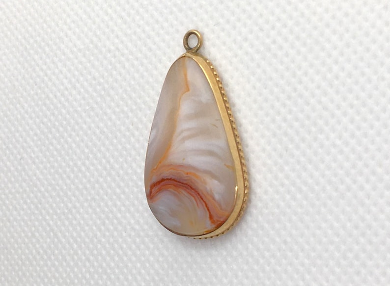 Peut inclure: Un pendentif en forme de larme avec une bordure dor&eacute;e et une pierre d'agate blanche et orange.