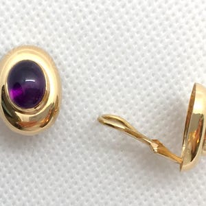 May include: A pair of gold-colored clip-on earrings. Each earring features an oval, deep purple gemstone set in a polished gold frame. The clip mechanism is visible on one earring.