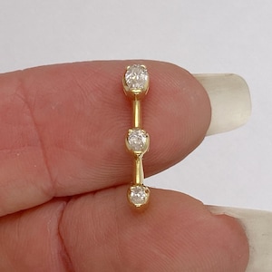 May include: A gold curved barbell body jewelry piece featuring three clear, round-cut stones set in prong settings along the length of the metal bar. The jewelry has a polished yellow gold finish and is designed for a piercing, showcasing a vertical alignment.