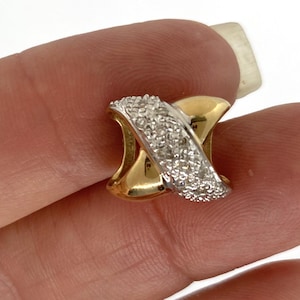 May include: A gold-toned ring with a crisscross design. One band is covered in small, sparkling crystals, while the other is smooth. The ring is held between fingers, showcasing its intricate details and elegant style.