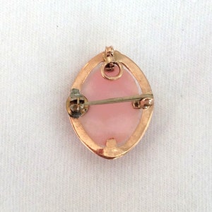 May include: A gold-toned oval brooch with a pink cameo set in the center. The brooch has a pin and catch closure on the back.