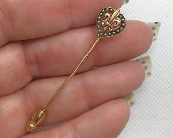 May include: A gold-toned stick pin with a heart-shaped head. The heart is adorned with small pearls and a fleur-de-lis design. The pin has a long, slender shaft and a decorative end.