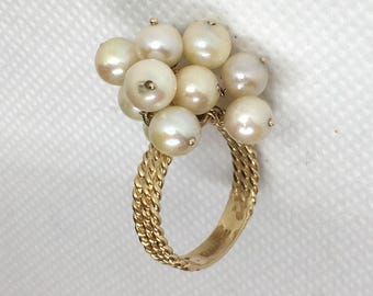 May include: A gold ring featuring a cluster of eleven white pearls. The ring's band has a braided design. The pearls are round and have a slight luster. The ring is set against a white background.