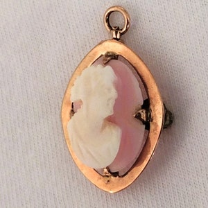 May include: A gold pendant with a pink cameo featuring a white profile of a woman. The cameo is set in a gold frame with a diamond-shaped design.