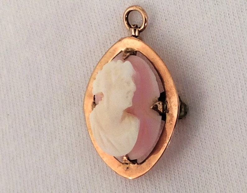 May include: A gold pendant with a pink cameo featuring a white profile of a woman. The cameo is set in a gold frame with a diamond-shaped design.