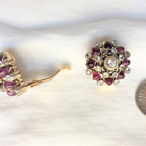 May include: Gold-toned clip-on earrings with a floral design. Each earring features a central clear stone surrounded by smaller clear stones and teardrop-shaped red stones. A US dime is included for size reference.