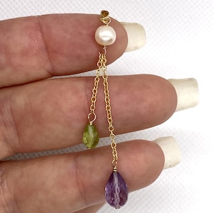 May include: A gold pendant featuring a white round pearl at the top, connected to two gold chains of different lengths. One chain ends in a small green teardrop-shaped gemstone, and the longer chain ends in a larger purple teardrop-shaped gemstone.