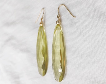 May include: A pair of long, faceted, light yellow-green earrings with gold-colored hooks. The earrings have a teardrop shape and are displayed against a white background.