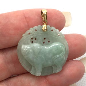 May include: A jade pendant with a gold bail. The pendant is carved with a light green elephant design within a circular frame. The elephant is standing, and the frame has a decorative top edge with small holes.