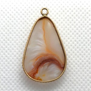 Peut inclure: Un pendentif en forme de larme avec un cadre en m&eacute;tal dor&eacute; et une pierre d'agate blanche avec des tourbillons bruns et orange.