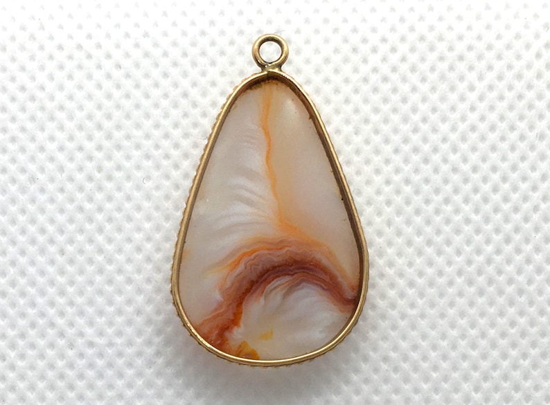 Peut inclure: Un pendentif en forme de larme avec un cadre en m&eacute;tal dor&eacute; et une pierre d'agate blanche avec des tourbillons bruns et orange.