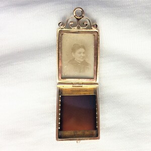 May include: A gold locket with a hinged front and a small photo of a woman inside. The locket is open and the inside is lined with a red fabric.