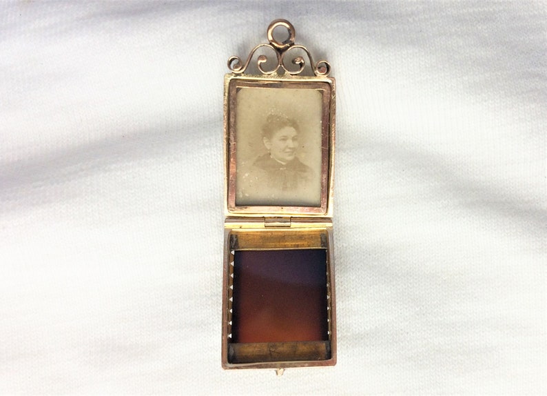 May include: A gold locket with a hinged front and a small photo of a woman inside. The locket is open and the inside is lined with a red fabric.
