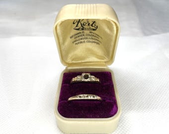 May include: An open, vintage jewelry box with two rings. The box is cream-colored with a gold satin interior and a purple velvet base. The rings are gold with small, clear stones. The top ring has a larger, dark stone.
