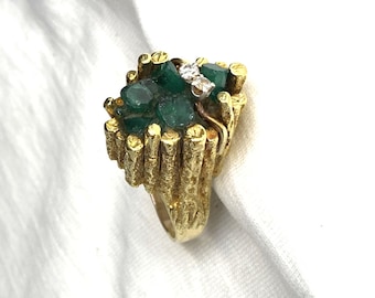 May include: A gold ring featuring a textured, bark-like band and a cluster of raw, deep green emeralds set atop the shank. Two small, clear, round-cut diamonds are embedded within the emerald cluster, adding a bright contrast to the dark stones.