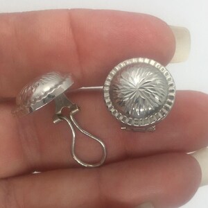 May include: A pair of silver-colored clip-on earrings. Each earring has a domed, circular design with a textured, radial pattern. The earrings are shown against a light background.