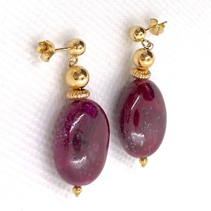 May include: A pair of gold earrings with a post-and-ball design. Each earring features a large, oval-shaped, deep red gemstone suspended below a gold bead and decorative element. The earrings have a polished finish.