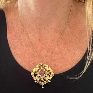 May include: A gold-toned pendant necklace featuring an intricate, openwork geometric design. The centerpiece is adorned with a central oval-cut pink gemstone, surrounded by small white pearls and dark accents, suspended on a thin gold-toned chain.