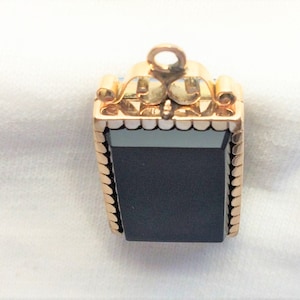 May include: A gold pendant with a black rectangular stone set in a gold frame. The frame has a decorative border of small gold beads.