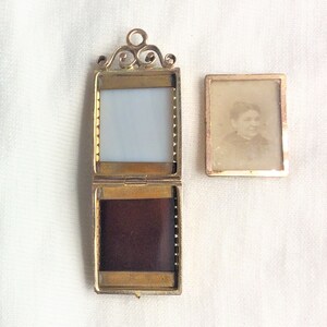 May include: A gold locket with a hinged front and back. The front has a clear glass window and the back has a dark velvet lining. The locket is open and a separate photo of a woman is visible.