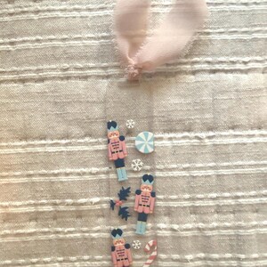 May include: A clear bookmark with Christmas-themed embellishments. Features three pink and black nutcracker soldiers, snowflakes, a candy cane, and a blue and white striped circle. A light pink ribbon is attached at the top.