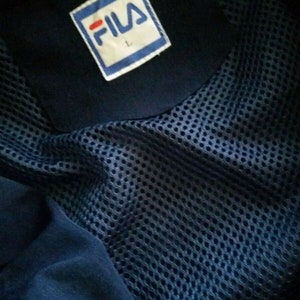 Puede incluir: Una chaqueta azul marino con forro de malla y un parche de logotipo Fila blanco en el pecho. El parche tiene una franja roja y la letra "L" impresa en &eacute;l.