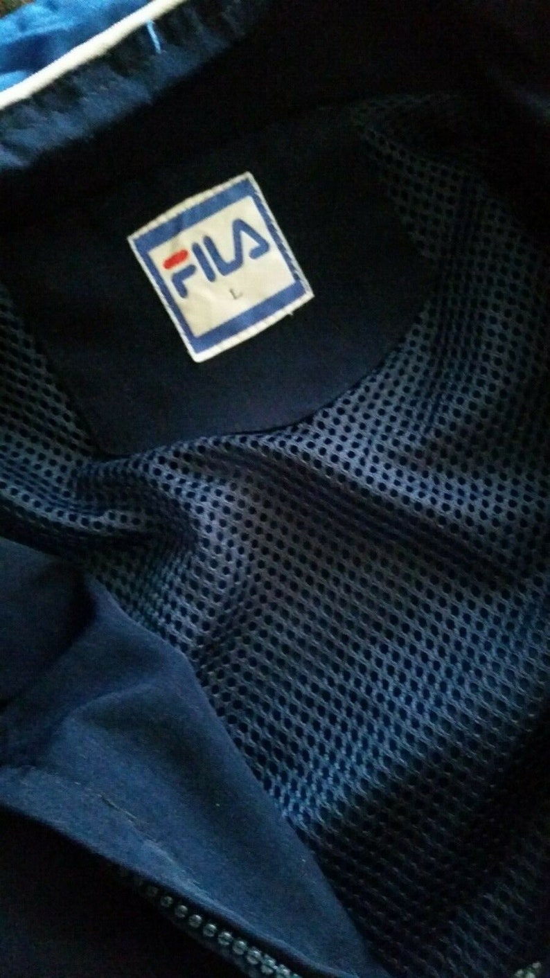 Puede incluir: Una chaqueta azul marino con forro de malla y un parche de logotipo Fila blanco en el pecho. El parche tiene una franja roja y la letra "L" impresa en &eacute;l.