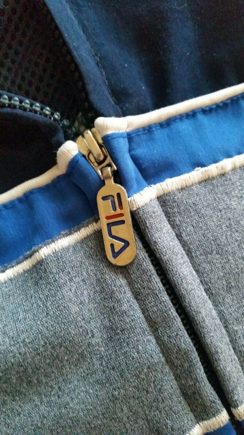 Puede incluir: Un primer plano de un tirador de cremallera con el logotipo de Fila. El tirador de cremallera es plateado con una franja roja y azul. El tirador de cremallera est&aacute; unido a una chaqueta a rayas azul y gris.