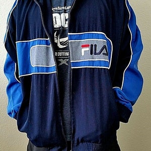 Puede incluir: Una chaqueta cortavientos azul marino y azul real con una franja blanca y gris en el pecho. La chaqueta tiene cierre de cremallera y un logotipo de Fila en el pecho.