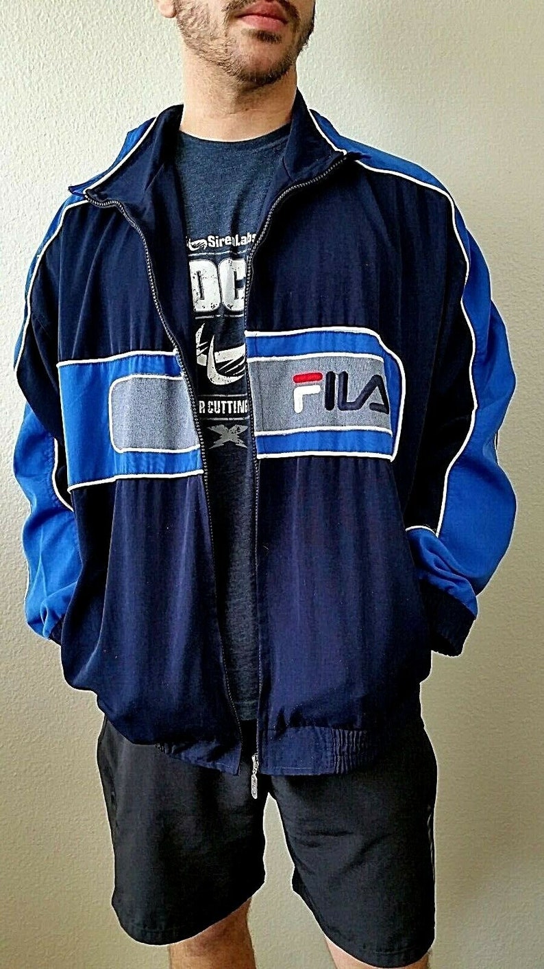 Puede incluir: Una chaqueta cortavientos azul marino y azul real con una franja blanca y gris en el pecho. La chaqueta tiene cierre de cremallera y un logotipo de Fila en el pecho.
