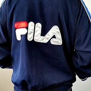 Puede incluir: Una chaqueta azul marino con rayas blancas y azules en las mangas. La chaqueta tiene un gran logotipo FILA blanco y plateado en la espalda.