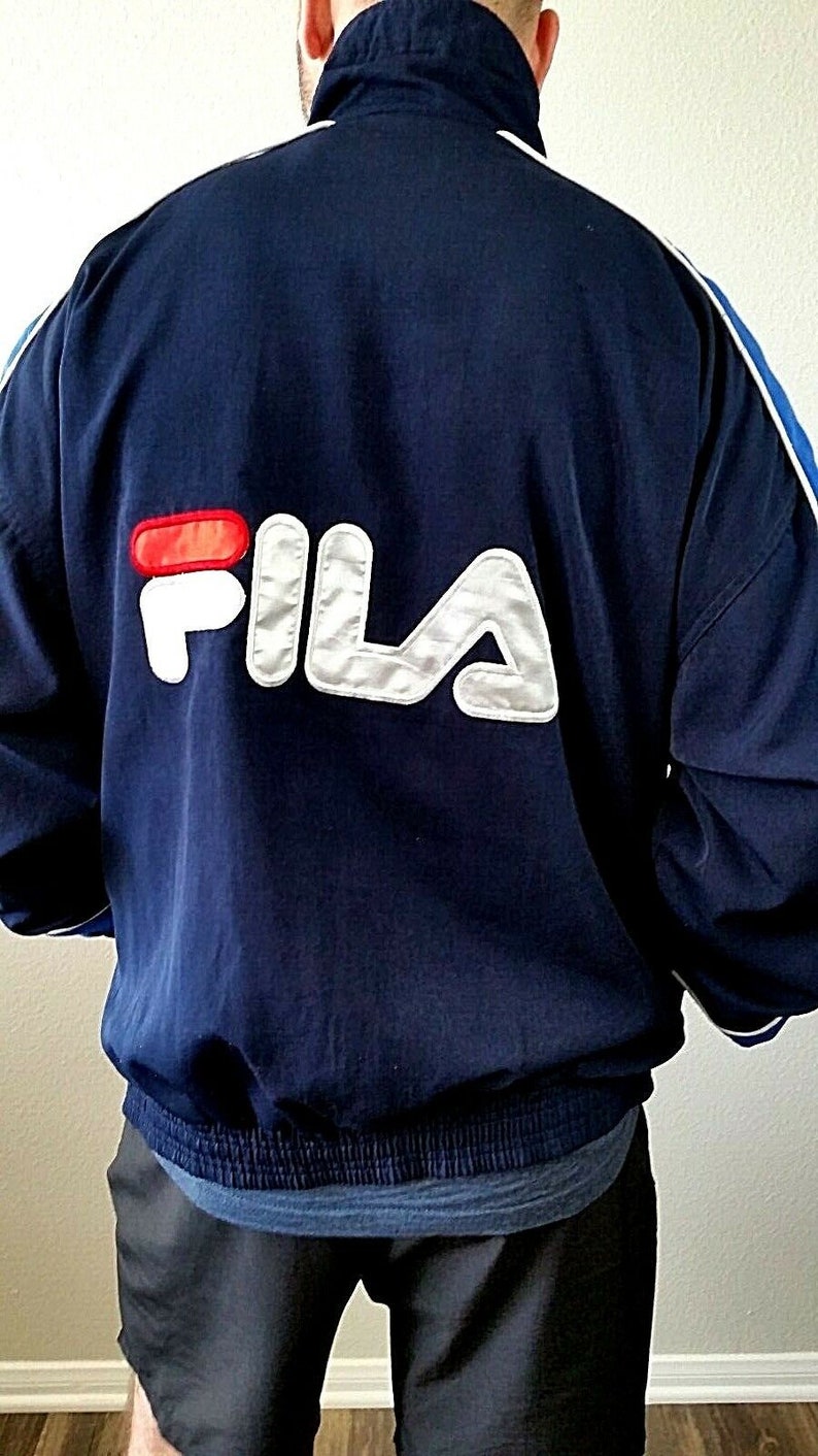 Puede incluir: Una chaqueta azul marino con rayas blancas y azules en las mangas. La chaqueta tiene un gran logotipo FILA blanco y plateado en la espalda.