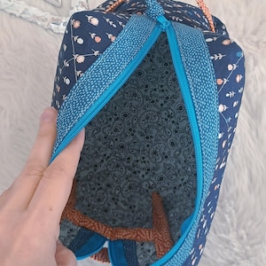 Puede incluir: Una bolsa de tela azul marino con un estampado floral y una cremallera turquesa. El interior presenta un diseño de cachemira. La bolsa tiene un ribete turquesa y un asa de cuerda naranja y azul.