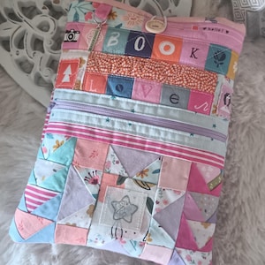 Puede incluir: Un bolso de libro patchwork en tonos rosa, azul y melocotón. El bolso presenta un cierre de cremallera, un parche en forma de estrella y la palabra "BOOK LOVER" escrita en cuadrados coloridos. Un pequeño dije cuelga de la cremallera.