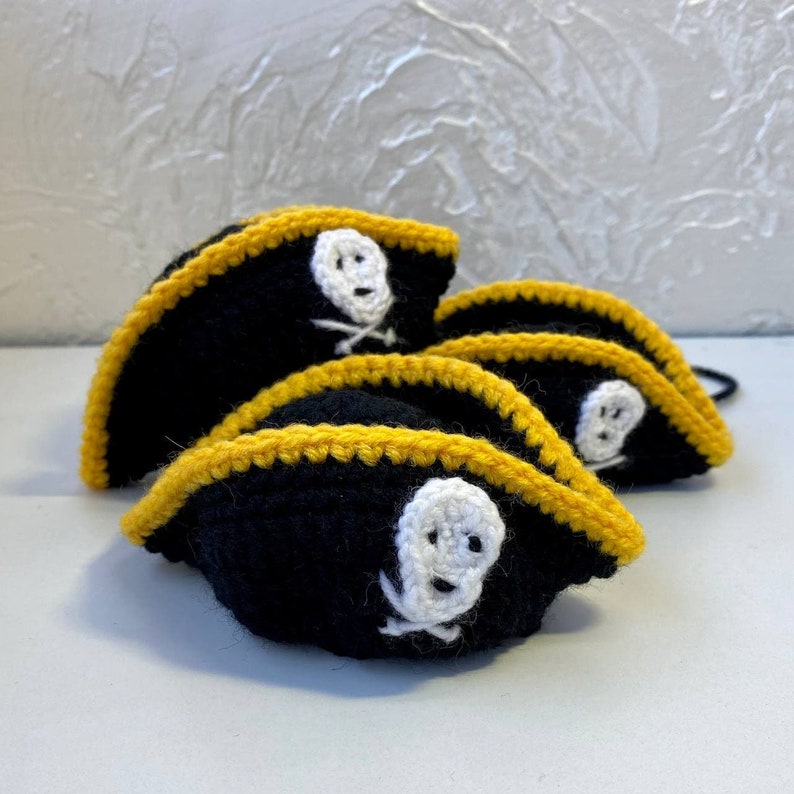 May include: Four crocheted pirate hats with black fabric and yellow trim. Each hat features a white skull and crossbones design.