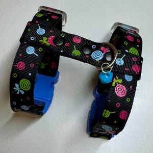 Puede incluir: Un arnés para perro negro con un patrón colorido de bastones de caramelo y espirales. El arnés tiene hebillas de plástico azules y un anillo plateado con una campanilla azul.