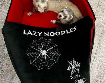 May include: Two ferrets in a red and black pet bed with the words "Lazy Noodles" embroidered on the front. The bed has a spider web design with a spider in the center and the word "Boo!" embroidered on the side.