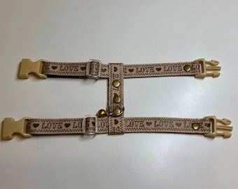 Puede incluir: Un arnés para gato de color beige con un patrón marrón de "LOVE" y herrajes de color dorado. El arnés tiene dos correas ajustables y un cierre de hebilla.