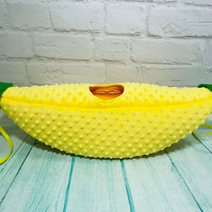 May include: A yellow banana-shaped plush toy with green fabric accents and a strap. The toy is made of soft, textured fabric and has a small opening at the top.